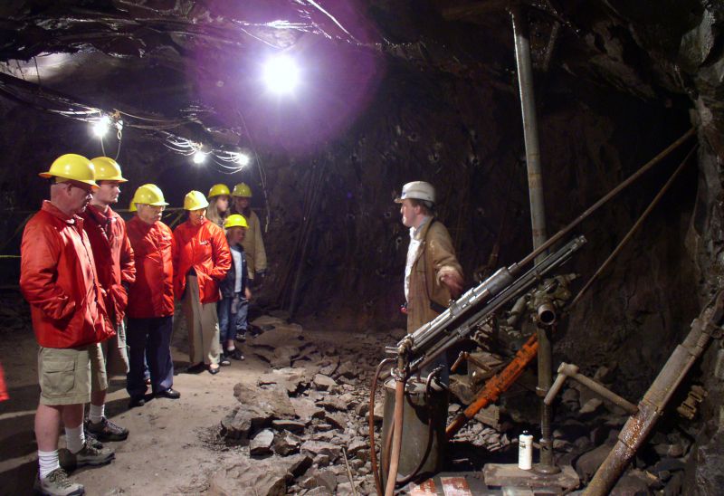 Tour group inside the quincy mine. The tour guide is on the right by a piece of drilling equipment. The tour group is wearing orange jackets, yellow hard hats and is listening to the guide.